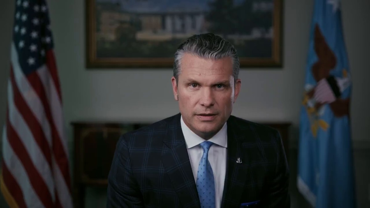 Pete Hegseth speaking to the camera in front of American and Department of War flags